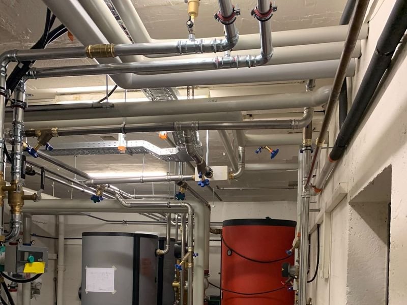 Plafond d'un local technique : réseau de conduites en inox serties avec vannes d'arrêt à poignée bleue, au-dessus d'un ballon gris et d'un ballon rouge.