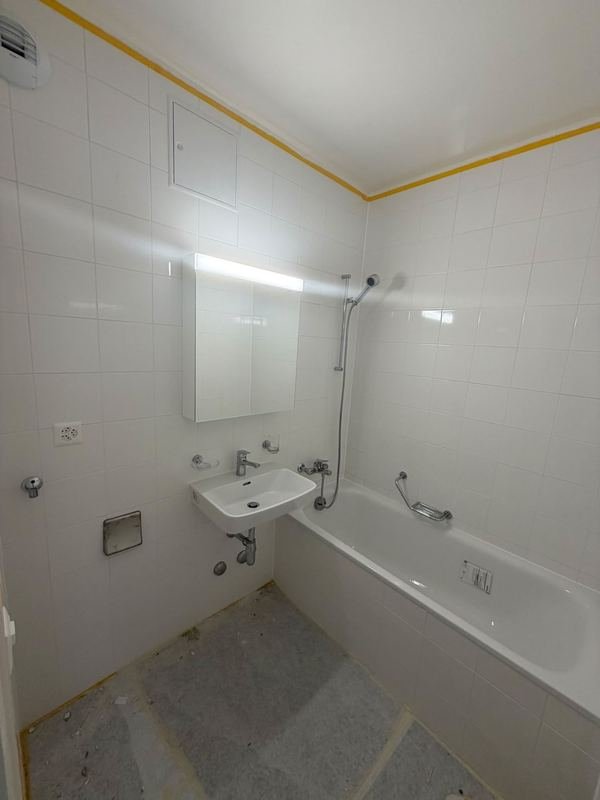 Salle de bains carrelée de blanc : baignoire avec barre de douche, lavabo, armoire à miroir éclairée.